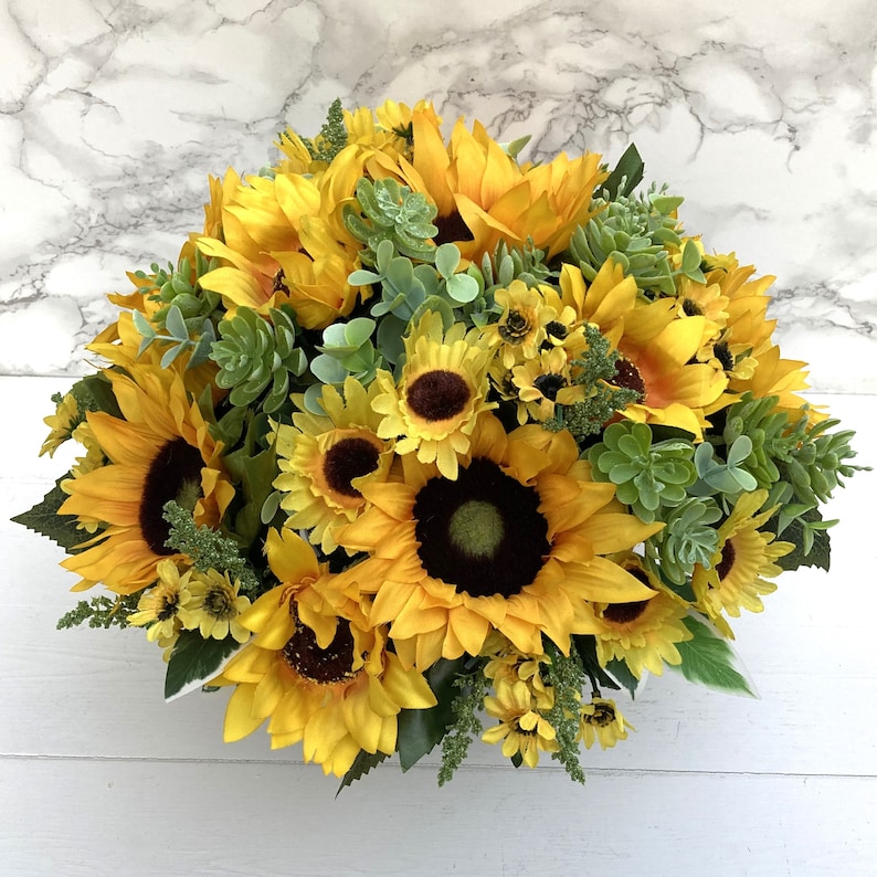 May include: A vibrant arrangement of sunflowers with bright yellow petals and dark brown centers. The bouquet includes green succulents and foliage, creating a full, textured display. The flowers are arranged in a round shape.