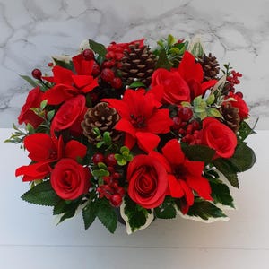 May include: A festive floral arrangement featuring vibrant red poinsettias, roses, and berries, accented with pinecones and lush green foliage. The arrangement is set in a dark container, creating a striking contrast. Perfect for holiday decor.