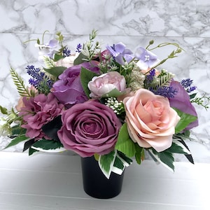 May include: A bouquet of artificial flowers in shades of pink, purple, and white arranged in a black vase. The flowers include roses, carnations, and lavender.