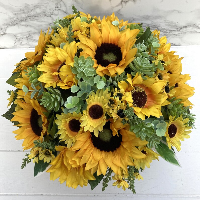 May include: A vibrant, full arrangement of sunflowers with dark brown centers and bright yellow petals. The arrangement includes green succulents and eucalyptus leaves, creating a natural, textured look. The flowers are clustered together, forming a dense, eye-catching display.