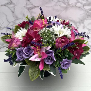 May include: A colourful floral display with purple, pink, and white blooms. The arrangement includes roses, lilies, and daisies, complemented by green leaves and lavender. The flowers are presented in a black container.