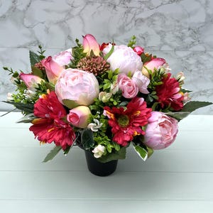 May include: A colourful floral display in a black pot. The arrangement includes pink peonies, red gerbera daisies, and green foliage. The flowers are arranged in a full, round shape, creating a lush and vibrant display.