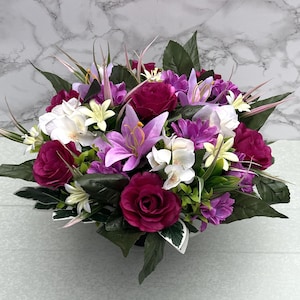 May include: A colourful floral arrangement with a variety of flowers in shades of purple, white, and burgundy. The arrangement includes roses, lilies, and other blooms, with green foliage. The flowers are arranged in a compact, rounded shape.