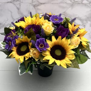 May include: A colourful floral arrangement with sunflowers, purple anemones, and yellow roses. The bouquet includes lavender sprigs and green foliage, arranged in a black pot. The sunflowers have dark brown centres and bright yellow petals.