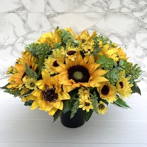 May include: A vibrant arrangement of sunflowers and greenery in a black pot. The sunflowers have bright yellow petals and dark brown centers. Green succulents and other foliage add texture and contrast to the floral display.