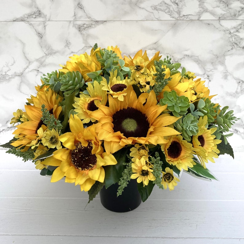 May include: A vibrant arrangement of sunflowers and greenery in a black pot. The sunflowers have bright yellow petals and dark brown centers. Green succulents and other foliage add texture and contrast to the floral display.