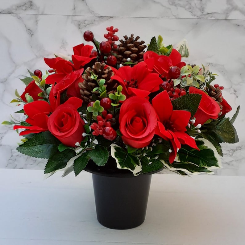 May include: A vibrant floral arrangement featuring red roses, poinsettias, berries, and pinecones. The arrangement is set in a black, cylindrical pot. Green leaves and accents provide contrast to the red flowers. A festive, decorative piece.