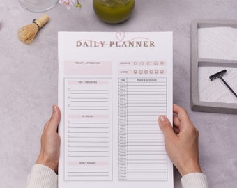 May include: A daily planner with sections for today's affirmation, top 5 priorities, to-do list, don't forget, mood, weather, and plans & schedule. The planner is open and held by a person's hands.