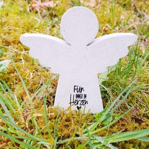 Könnte beinhalten: Eine weiße Beton-Engelfigur mit Flügeln, die auf einem Bett aus grünem Gras steht. Der Engel trägt die Inschrift "Für immer im Herzen".