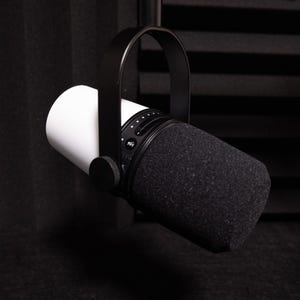 May include: A professional-grade microphone with a white cylindrical body and a black, foam-covered head. The microphone is suspended by a black, curved arm and is designed for recording or broadcasting.