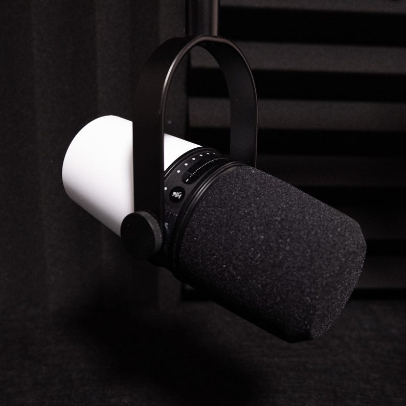May include: A professional-grade microphone with a white cylindrical body and a black, foam-covered head. The microphone is suspended by a black, curved arm and is designed for recording or broadcasting.