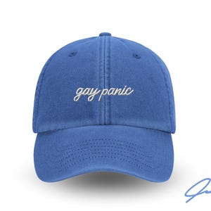 Può includere: Un cappellino da baseball blu con la scritta ricamata "gay panic" in corsivo bianco. Il cappellino ha una visiera curva e un classico design a sei pannelli. Una firma è visibile in basso a destra.