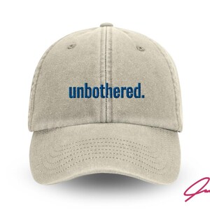 Op de afbeelding: Beige baseballpet met het woord "unbothered" in blauw geborduurd. De pet heeft een gebogen klep en een gestikt ontwerp. Een handtekening staat in de rechter benedenhoek.