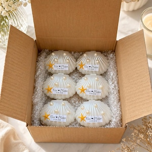 May include: A brown cardboard box containing six white seashell-shaped candles. Each candle is decorated with small white pearls and a small orange starfish. A white label on each candle features the text Nici heart Matze and 23.05.2026.