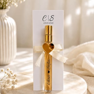 Può includere: Un flacone di profumo in vetro trasparente con tappo e pompetta dorati, fissato su un cartoncino rettangolare bianco. Il cartoncino riporta le iniziali "e|S" e la data "08.09.2026". Un nastro color crema con un ciondolo a forma di cuore dorato è legato attorno al flacone, su cui sono stampati i nomi "Chris" e "Sofia".
