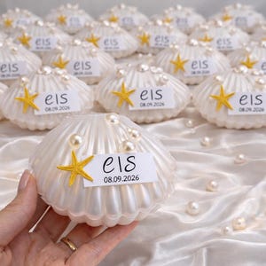 Puede incluir: Un adorno de concha marina blanca y estriada, decorado con una pequeña estrella de mar amarilla y varias perlas blancas. Lleva una etiqueta rectangular blanca en el frente con el texto "els" en letra cursiva sobre la fecha "08.09.2026".