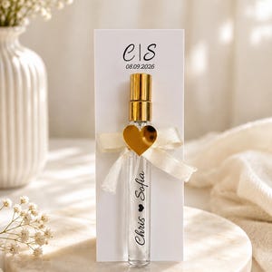 Può includere: Un flacone di profumo in vetro trasparente con tappo spray dorato, fissato su un cartoncino rettangolare bianco. Il cartoncino riporta il testo "e|S" e "08.09.2026". Il flacone è decorato con un nastro color crema e un ciondolo a cuore dorato, con "Chris ❤ Sofia" stampato sul vetro.