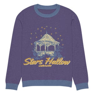 May include: A purple sweatshirt with a blue collar and cuffs. The front features a gazebo design with the words "Stars Hollow" in gold lettering, surrounded by yellow stars. A cozy, long-sleeved top.
