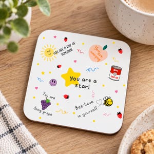 May include: A square white coaster featuring colourful illustrations including a yellow sun, a peach, a soup can, grapes, and a bee. Text includes: You are a ray of sunshine, You are amazing, You are a star!, You are doing grape, and Bee-lieve in yourself.
