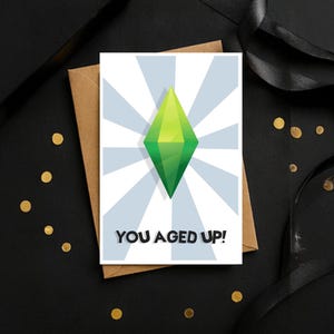 May include: A rectangular greeting card featuring a white background with light blue radiating sunburst stripes. Centered is a bright green diamond-shaped plumbob icon. Below the icon, the text reads YOU AGED UP! in bold black capital letters.