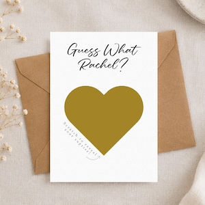 May include: A white card with the text "Guess What Rachel?" and a gold heart-shaped scratch-off area. The card is placed in a brown envelope, with dried flowers in the background. The text "Scratch to reveal your surprise!" is also on the card.
