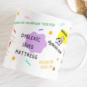 May include: A white ceramic mug featuring a purple blob with the text Dyslexic Lives Mattress, alongside a small cow illustration labelled Dyslexic cow, the text Yuck Fou, and other colourful doodles including stars, squiggles, and a small poop emoji.