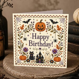 May include: A square greeting card featuring a cross-stitch design with the text Happy Birthday. The illustration includes a jack-o'-lantern, a black cat, a ghost, a haunted house, bats, spiders, autumn leaves, and purple flowers on a cream background.