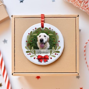 May include: A round white ceramic ornament featuring a photograph of a golden retriever dog with its tongue out. The dog is framed by a green leafy wreath with red berries and a red bow at the bottom. The name Archie is printed in black script.