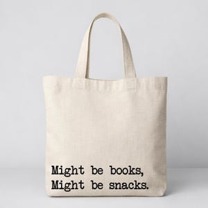 May include: A cream-coloured canvas tote bag featuring the text "Might be books, Might be snacks" in black. The bag has two handles for carrying. The design is simple and practical, suitable for carrying books, snacks, or other items.