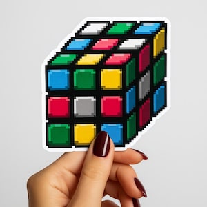 May include: A pixel art style sticker of a 3D cube puzzle featuring a grid of small squares in various colors including red, blue, green, yellow, white, and gray, all outlined in black, presented as a die-cut sticker with a white border.