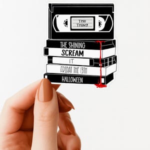 May include: A black and white sticker featuring a stack of VHS tapes with horror movie titles: "The Thing," "The Shining," "Scream," "It," "Friday the 13th," and "Halloween." Red blood drips down the side. The sticker is held by a hand.