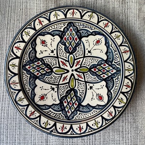 Può includere: Un piatto in ceramica bianca con un motivo geometrico blu, verde e rosso. Il piatto ha un bordo ondulato ed è decorato con dettagli intricati.