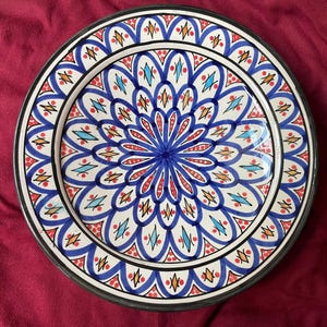 Op de afbeelding: Een wit keramiek bord met een blauw, rood en oranje geometrisch patroon. Het bord heeft een verhoogde rand en een glanzende afwerking.