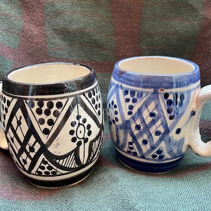 May include: Two ceramic mugs with geometric patterns. One mug is black and white with a geometric pattern of black dots and lines. The other mug is blue and white with a geometric pattern of blue lines and dots.