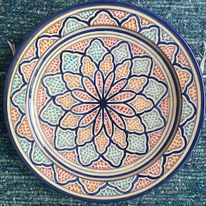 Può includere: Un piatto in ceramica decorativo con un motivo geometrico blu, bianco, arancione e rosa. Il piatto ha un bordo rialzato e un disegno centrale che ricorda un fiore.