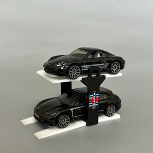 Puede incluir: Un elevador de coches de juguete negro con dos coches Porsche de juguete negros encima. El coche de arriba tiene una franja blanca y el coche de abajo tiene un logotipo de Martini Racing.