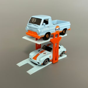 Puede incluir: Un elevador de coches de juguete blanco con dos coches de juguete en &eacute;l. El coche de arriba es una camioneta Dodge azul claro con un logotipo de Gulf en el lateral. El coche de abajo es un Porsche blanco con rayas de carreras naranja y negras y el n&uacute;mero 68 en el lateral.