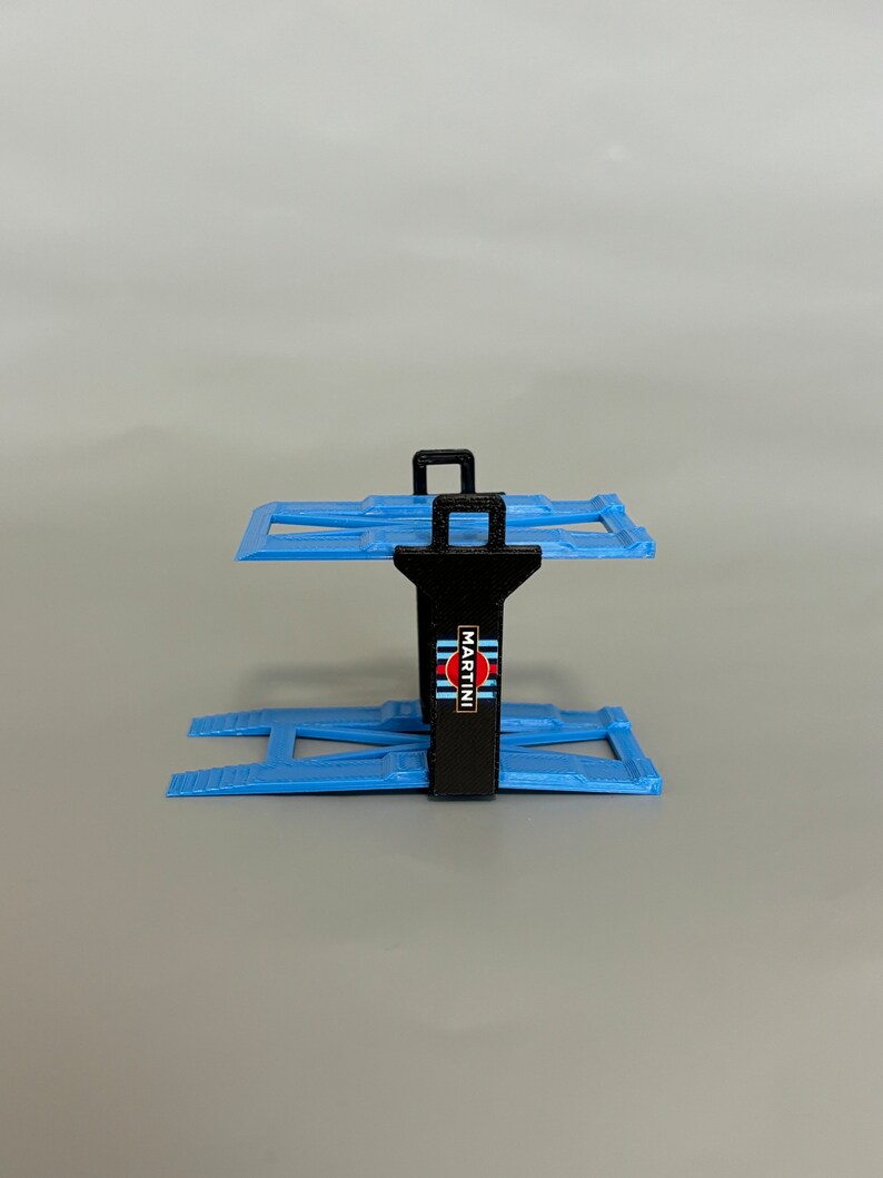 Puede incluir: Un elevador de coches de pl&aacute;stico negro y azul con un logotipo de Martini Racing. El elevador est&aacute; dise&ntilde;ado para sujetar coches de juguete.