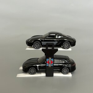 Puede incluir: Dos coches de juguete Porsche negros en una rampa blanca con un elevador negro. El coche de arriba tiene un logotipo de Martini Racing en el lateral.