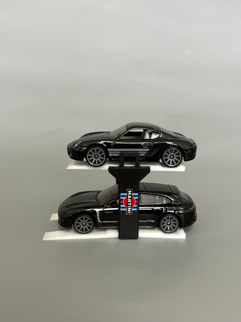 Puede incluir: Dos coches de juguete Porsche negros en una rampa blanca con un elevador negro. El coche de arriba tiene un logotipo de Martini Racing en el lateral.