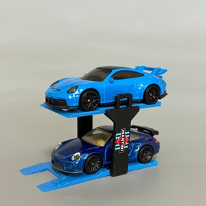Puede incluir: Un elevador de coches de juguete azul con dos coches Porsche de juguete azules. El coche de arriba tiene un techo negro y el coche de abajo tiene un logotipo de Martini Racing en el lateral.