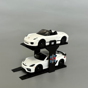 Puede incluir: Un elevador de coches negro de dos niveles con un Porsche 911 GT3 RS blanco con la decoraci&oacute;n de Martini Racing en el nivel inferior y un Porsche Boxster Spyder blanco en el nivel superior.