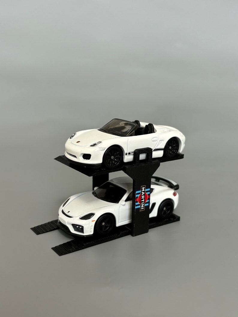 Puede incluir: Un elevador de coches negro de dos niveles con un Porsche 911 GT3 RS blanco con la decoraci&oacute;n de Martini Racing en el nivel inferior y un Porsche Boxster Spyder blanco en el nivel superior.