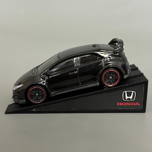 Puede incluir: Un coche modelo negro Honda Civic Type R con ruedas con detalles rojos se muestra en una rampa negra. El logotipo de Honda es visible en la rampa. El coche tiene un alerón y un diseño elegante.