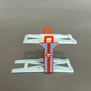 Puede incluir: Un elevador de coches de pl&aacute;stico blanco con detalles naranjas y un logotipo de Gulf. El elevador est&aacute; hecho de dos rampas y un soporte central con un asa.