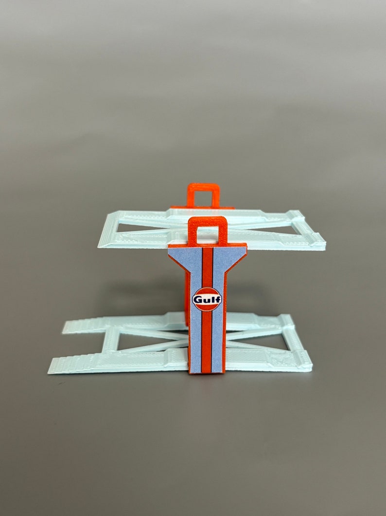 Puede incluir: Un elevador de coches de pl&aacute;stico blanco con detalles naranjas y un logotipo de Gulf. El elevador est&aacute; hecho de dos rampas y un soporte central con un asa.