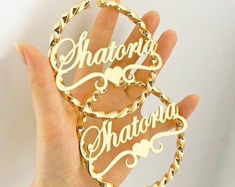 Könnte beinhalten: Goldene Creolen mit dem Namen "Shatoria" in kursivschrift mit einem kleinen Herz-Anhänger.