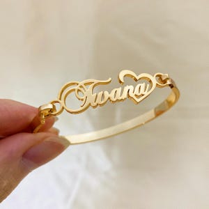 May include: Gold-toned bangle bracelet featuring the name "Juana" in a cursive script with a heart charm.