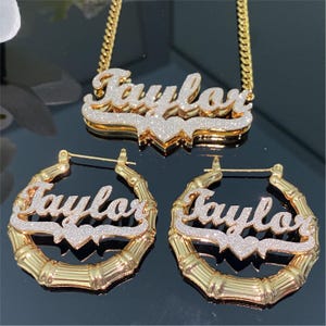 May include: A gold-colored jewelry set featuring the name "Taylor". The set includes a necklace with a chain, and a pair of hoop earrings. The name is in a cursive font with a glittery, silver-colored fill. The earrings have a bamboo design.