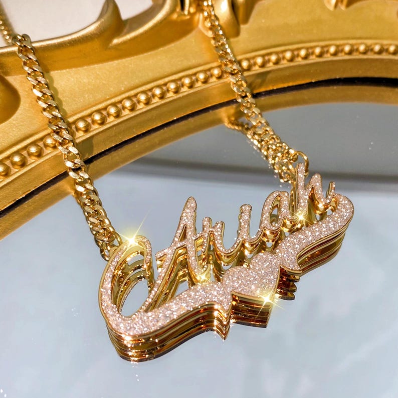 K&ouml;nnte beinhalten: Eine goldene Kettenkette mit einem mehrschichtigen, glitzernden Namensschild-Anh&auml;nger, auf dem "Ariah" steht.